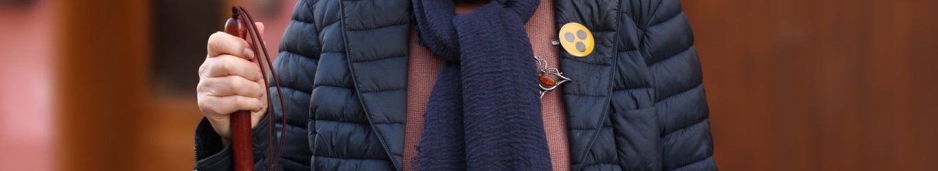 Person mit Langstock Man sieht eine Person in einer blauen Steppjacke mit einem Blinden-Symbol-Buttton. In der Hand hält sie einen Langstock.